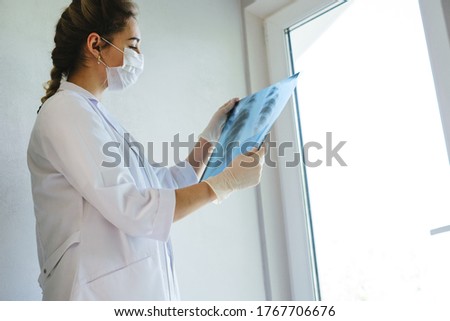 A radiologist in a medical mask and gloves radiates an x-ray image of the patient's lungs. The girl doctor works on the front line. The doctor is holding an X-ray of his lungs.