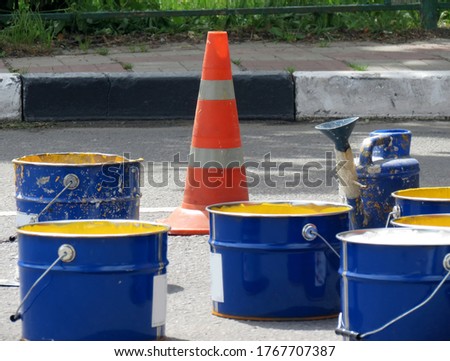 Repair work on the road. Blue large cans with white and yellow paint, next to it is an orange traffic signal cone                               