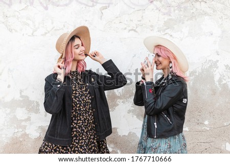 Fashion young woman photographer takes picture happy girl with sweet smile with pink hair in casual denim jacket that straightens vintage hat. Stylish sisters take photo on modern camera near wall.