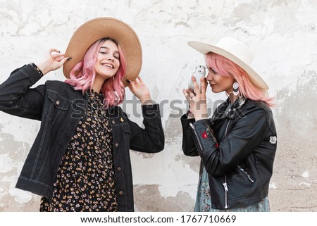 Stylish woman with pink hair in youth fashion clothes takes pictures on camera of joyful pink-haired girl with positive smile in trendy elegant clothes. Fashionable friends take snapshots on street.