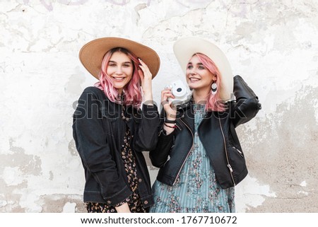 Happy woman photographer with lovely smile with pink hair in leather jacket in hat with modern camera poses next to smiling pink-haired girlfriend in fashion denim jacket near vintage wall on street.