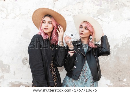 Stylish young woman sisters in fashionable summer youth clothes in trendy straw hats posing near vintage wall. Lovely urban girl in blue dress stands with modern camera near cute girlfriend in city.