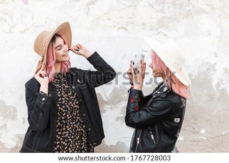 Fashion woman with pink hair in youth casual clothes takes photo on camera of happy pink-haired girl with positive smile in trendy elegant clothes. Pretty fashionable friends take snapshots outdoors.