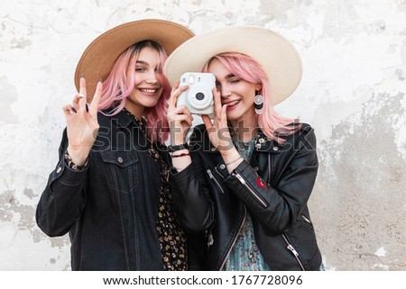 Happy pink-haired girl with smile in straw hat in fashion jacket stands next to girlfriend with modern camera in jacket and shows gesture of peace. Attractive funny girls take positive pictures.
