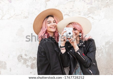 Smiling stylish young woman sisters in fashion summer youth clothes in trendy straw hats posing near vintage wall.Happy cute girl in blue dress stands with modern camera near cute girlfriend in street