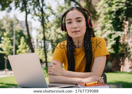 Portrait of pensive asian woman wearing red headphones, looking at camera. Young beautiful girl using laptop, studying, distance learning, online education concept  