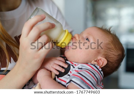 Mother holds her two moths old baby on the chair taking care at home - Caucasian woman with her newborn child bottle feeding milk on lap - Affectionate and bonding childhood and motherhood concept Royalty-Free Stock Photo #1767749603
