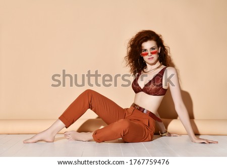 Beautiful young slim red haired woman in trousers, bra and sunglasses sitting in relaxed pose and looking at camera over pastel yellow background. Stylish women looks concept