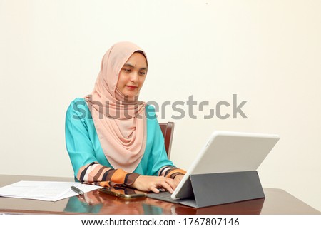 young Asian Malay Muslim woman wearing headscarf at home office student sitting at table talk mingle look at computer pone book document study smile happy look at computer type