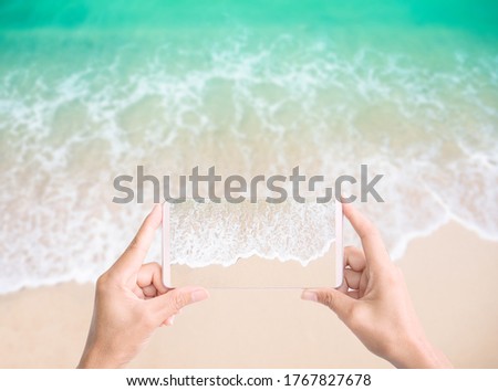 Hand woman holding mobile with sand beach view on blur image of sand beach with white sea soft wave on top view . 