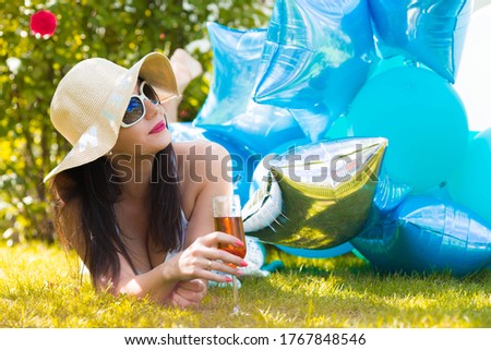 A young girl in sunglasses on her birthday is lying on a green lawn with a glass of wine and balloons in the background. The concept of a holiday and a good mood.