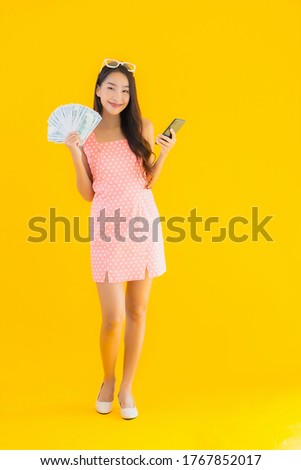 Portrait beautiful young asian woman show a lot of cash and money with mobile smart phone on yellow isolated background