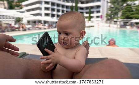A cute baby playing with a cell phone at a swimming. In his father's lap