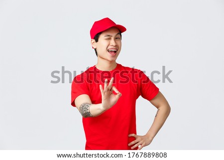 Best deal on market. Handsome smiling asian delivery guy in red cap and t-shirt ensure best quality of delivering goods, safety of parcels. Courier show okay, approval sign, wink cheeky camera