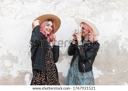 Happy two young girls in fashionable clothes travel and take pictures with a modern camera near a white wall on the street. Girlfriends have a good time
