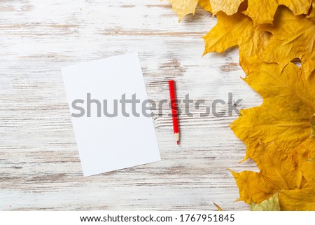 White paper sheet and pencil lies on vintage wooden desk with bright foliage. Flat lay composition with autumn leaves on white wooden surface. Blank notepaper with copy space for design.