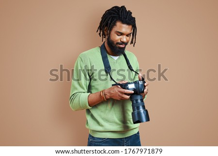 Handsome young African photographer in casual clothing using digital camera and smiling while standing against brown background