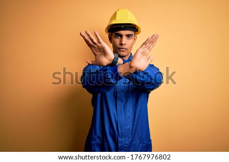 Young african american worker man wearing blue uniform and security helmet Rejection expression crossing arms and palms doing negative sign, angry face