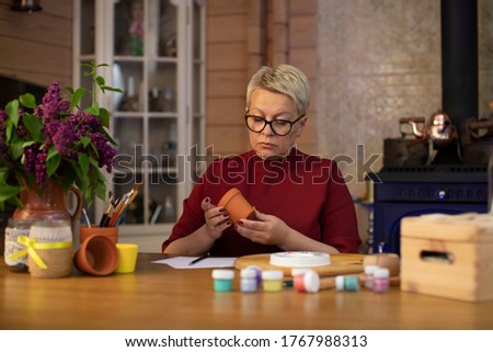 attractive middle-aged woman coming up with drawing for a flowerpot in cozy country house with fireplace Concentrated woman with blond hair dressed casually painting