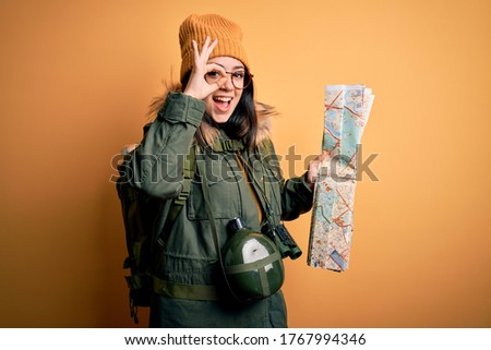 Young hiker woman wearing hiking backpack looking at tourist map over yellow background with happy face smiling doing ok sign with hand on eye looking through fingers