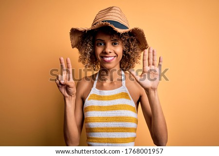 African american tourist woman with curly on vacation wearing summer hat and striped t-shirt showing and pointing up with fingers number seven while smiling confident and happy.