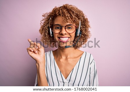 African american curly call center agent woman working using headset over pink background with a big smile on face, pointing with hand and finger to the side looking at the camera.