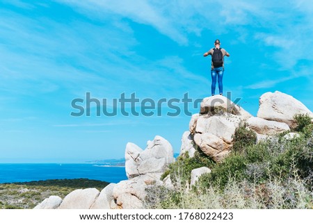 a young caucasian man, seen from behind, wearing jeans and a t-shirt and carrying a backpack, on the top of a rock formation, taking a photo of the sea in the Southern coast of Corsica, in France