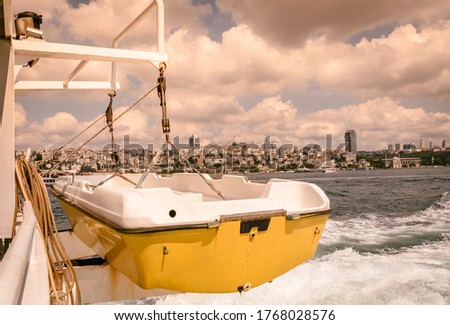 small yellow-white boat hanging from a moving ship, large white clouds, foaming sea and the city in behind as background view.