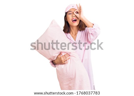 Young beautiful chinese girl wearing sleep mask and pajama holding pillow smiling happy doing ok sign with hand on eye looking through fingers 