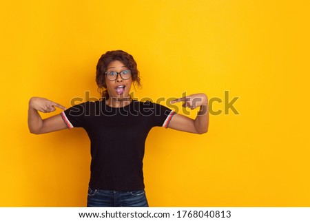 Pointing at herself. African-american young woman's portrait isolated on yellow studio background. Beautiful female curly model. Concept of human emotions, facial expression, sales, ad, youth.