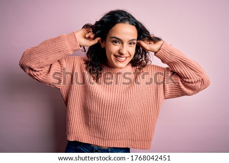 Young beautiful woman with curly hair wearing casual sweater over isolated pink background Smiling pulling ears with fingers, funny gesture. Audition problem