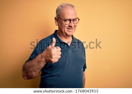 Middle age handsome hoary man wearing casual polo and glasses over yellow background doing happy thumbs up gesture with hand. Approving expression looking at the camera showing success.