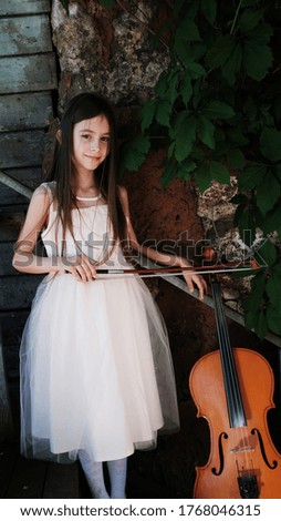 beautiful girl in a pink dress stands with a cello in a country house with wild grapes