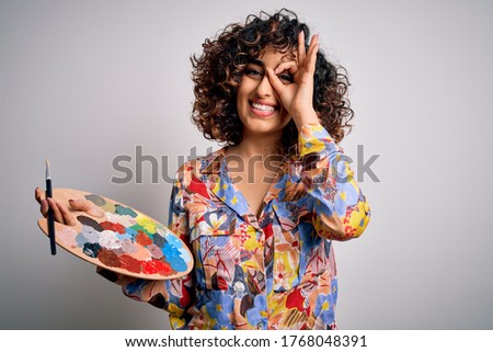 Young beautiful curly arab artist woman painting using brush and palette with colors with happy face smiling doing ok sign with hand on eye looking through fingers