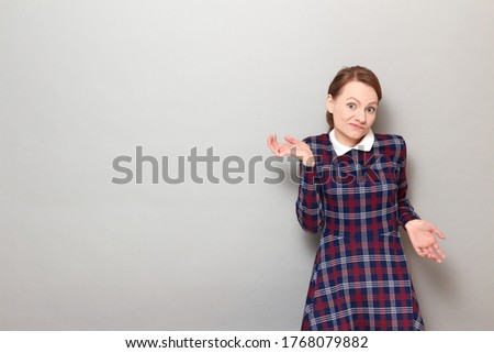 Studio portrait of funny confused blond girl wearing checkered dress, grimacing, waving hands, shrugging shoulders, not understanding how this happened, standing over gray background, with copy space