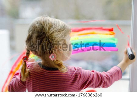 Adoralbe little toddler girl with rainbow painted with colorful window color during pandemic coronavirus quarantine. Child painting rainbows and hearts around the world with words Let's all be well.