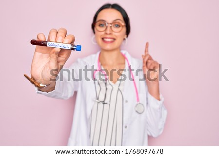 Beautiful doctor woman with blue eyes wearing stethoscope holding coronavirus test tube surprised with an idea or question pointing finger with happy face, number one