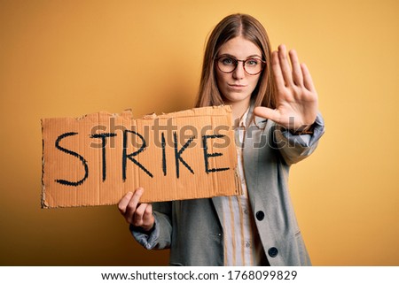 Young beautiful redhead woman holding banner with strike message over yellow background with open hand doing stop sign with serious and confident expression, defense gesture