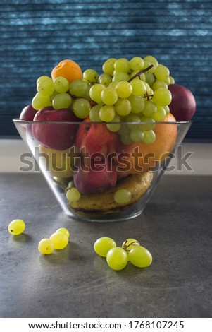 Fresh summer fruits in a glass vase. Colorful still life with grapes, mango, bananas, pears, peaches, nectarines and apricots on grey table. Navy blue background. 