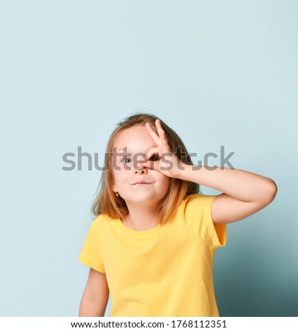 Adorable blonde little girl in yellow t-shirt. She is showing ok sign and looking through it while posing against turquoise studio background. Childhood, gestures, emotions. Close up, copy space