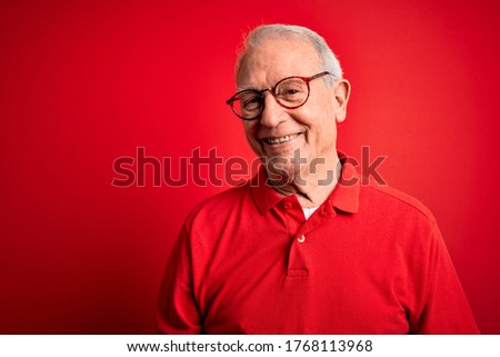 Grey haired senior man wearing glasses and casual t-shirt over red background Relaxed with serious expression on face. Simple and natural looking at the camera.