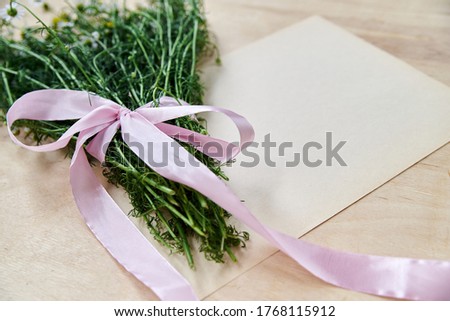 Small camomiles bouquet tied with pink ribbon and white paper sheet on wooden table. Natural background picture of wild flowers. Ecological texture for poems, letters, romantic notes.