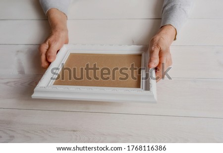 Elderly woman looking at the picture, light wodden background. Senior wrinkled hands holding white photo frame.  Empty template, mock-up. Happy memories, sadness, nostalgia concept.