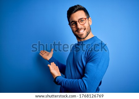 Young handsome man with beard wearing casual sweater and glasses over blue background Inviting to enter smiling natural with open hand Royalty-Free Stock Photo #1768126769