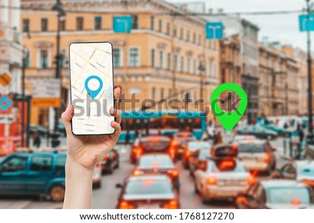 A female's hand holds a smartphone with an online map app. In the background is a city street with traffic jam and green location icon.Concept of online navigation and GPS