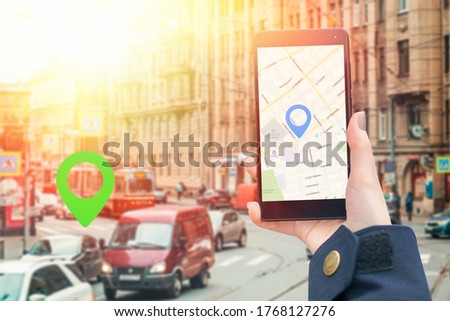 A woman's hand in a jacket holds a smartphone with an online map app. In the background is a blurred road with a cars and icon of location.The concept of online navigation and modern technologies