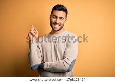 Young handsome man wearing casual sweater standing over isolated yellow background with a big smile on face, pointing with hand and finger to the side looking at the camera.