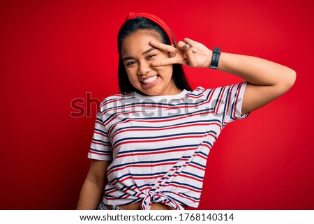 Young beautiful asian girl wearing casual striped t-shirt over isolated red background Doing peace symbol with fingers over face, smiling cheerful showing victory