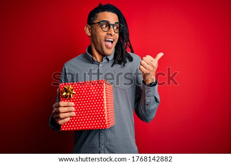 African American man with dreadlocks holding present box over red isolated background pointing and showing with thumb up to the side with happy face smiling