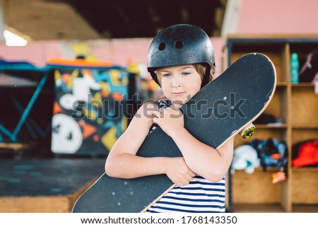 Portrait of handsome caucasian boy athlete skateboarder in protective helmet with skateboard in hands looking at camera on background of skate park. A child and an active hobby, sports and health.